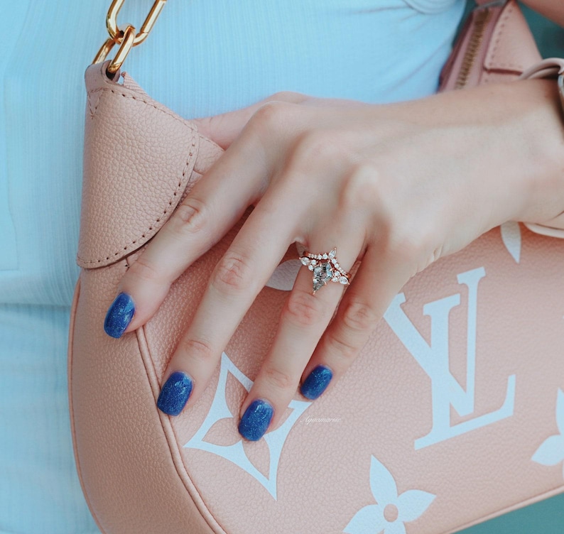 May include: A close-up of a hand wearing a pear-shaped diamond engagement ring with a delicate band. The ring is set in a rose gold band and features a halo of smaller diamonds. The hand is resting on a pink leather handbag with a white LV logo.