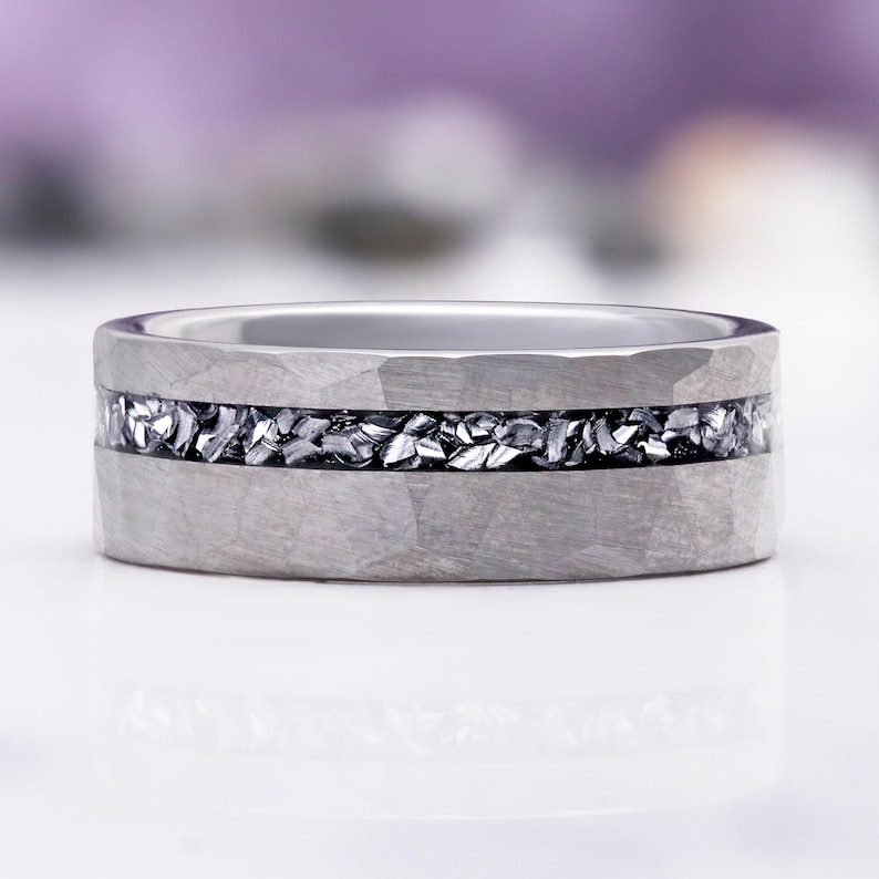 Pu&ograve; includere: Un anello color argento caratterizzato da una finitura in metallo martellato sulla fascia esterna. Un inserto orizzontale di frammenti scuri dall'aspetto metallico attraversa il centro della fascia, creando un contrasto con la superficie.