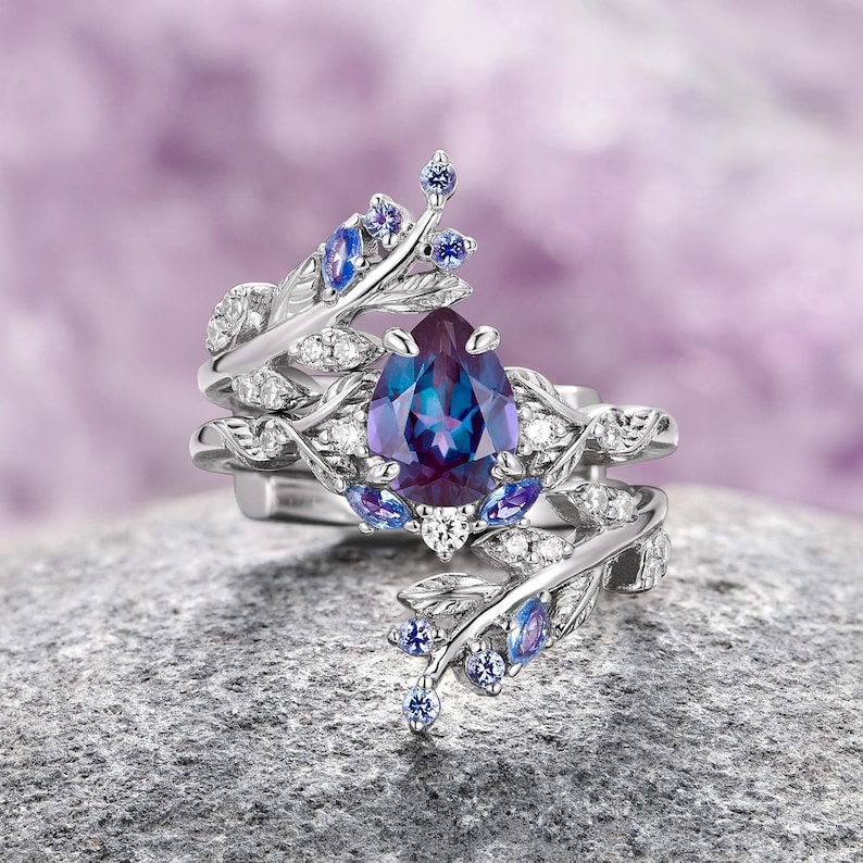 Op de afbeelding: Een ring met een peervormige, paarse en blauwe edelsteen, een zilveren band en ingewikkelde bladachtige details. De ring is bezet met kleinere blauwe edelstenen en witte diamanten.