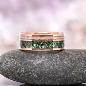 May include: A rose gold band ring featuring two distinct inlays. The top inlay consists of a shimmering, iridescent opal material, while the bottom inlay is a textured, deep green moss agate stone. The word Aquamarise is printed on the band.