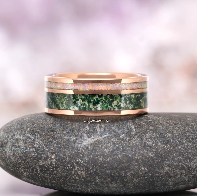 May include: A rose gold band ring featuring two distinct inlays. The top inlay consists of a shimmering, iridescent opal material, while the bottom inlay is a textured, deep green moss agate stone. The word Aquamarise is printed on the band.