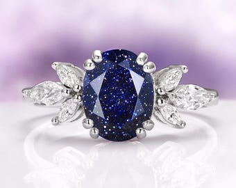 May include: A silver ring featuring a large, oval, dark blue gemstone with a sparkling appearance. The ring is accented with smaller, clear, marquise-cut stones on each side, creating a floral design. The ring is set against a soft purple background.