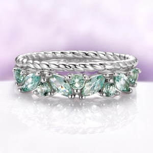 May include: Two silver rings. The top ring has a twisted rope design. The bottom ring features a series of light green, marquise-cut gemstones. The gemstones are set in a silver band, creating a floral-like pattern. The background is a soft purple.