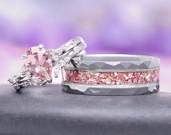 May include: A pair of wedding rings. One ring features a pear-shaped pink gemstone with surrounding accent stones. The other ring is gray with a pink crushed stone inlay. The rings are set against a blurred purple background.