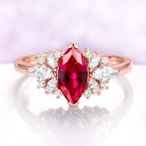May include: Rose gold ring featuring a large, marquise-cut ruby center stone. The ruby is surrounded by smaller, white, pear-shaped and round accent stones. The ring's band is a polished rose gold.