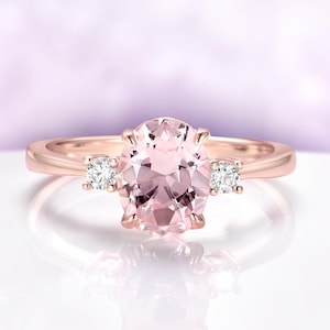May include: A rose gold ring featuring a large, oval-cut, light pink gemstone flanked by two smaller, round, clear gemstones. The ring has a polished finish and is set against a soft, blurred background.