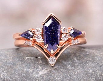 Puede incluir: Un anillo de oro rosa con una piedra central hexagonal de arenisca azul, flanqueada por dos piedras laterales en forma de cometa y pequeños acentos redondos transparentes. La banda tiene forma de chevrón y el texto Aquamarise es visible.