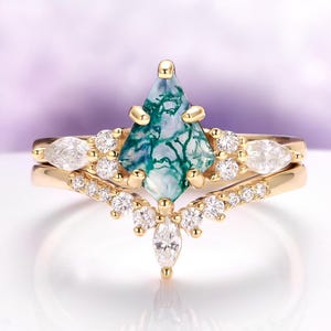 Könnte beinhalten: Ein goldener Ringsatz mit einem zentralen, rautenförmigen grünen und blauen Edelstein. Der Ring ist mit kleineren, klaren Edelsteinen und einem zarten Band verziert. Das Set enthält ein passendes Band mit zusätzlichen klaren Edelsteinen.