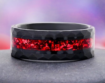May include: A black ring with a hammered, faceted texture on the outer edges and a wide, horizontal inlay of vibrant, crushed red material running through the center. The band has a smooth, dark interior and a polished, reflective finish.