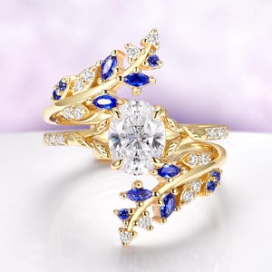 May include: A gold-toned ring set with an oval, clear gemstone and accented with blue and white gemstones in a leaf-like design. The ring has a split shank and is set on a white surface.
