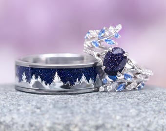 Puede incluir: Un par de alianzas de plata con piedras preciosas azules y blancas. La banda de la izquierda presenta un diseño de un bosque con árboles. La banda de la derecha presenta una piedra preciosa azul rodeada por un diseño de vid de plata con piedras preciosas azules y blancas.