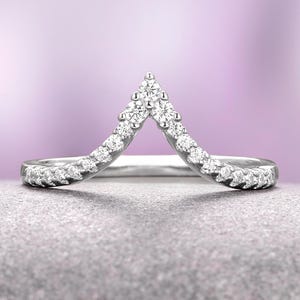 May include: A silver-toned ring with a chevron design, set with multiple small, clear gemstones. The ring's band curves upward to form a V-shape, with the gemstones arranged along the band and at the peak of the V.