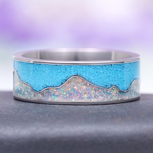 May include: A silver ring with a mountain range design. The top section is filled with small, light blue stones, while the bottom section features iridescent stones. The ring has a polished, metallic finish.