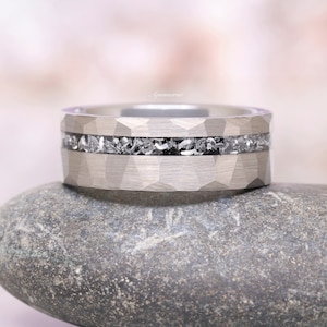 Pu&ograve; includere: Un anello in metallo color argento con una superficie esterna sfaccettata e martellata e un intarsio orizzontale centrale riempito di frammenti scuri, testurizzati e dall'aspetto metallico. La parola Aquamarise &egrave; scritta in piccolo corsivo chiaro.