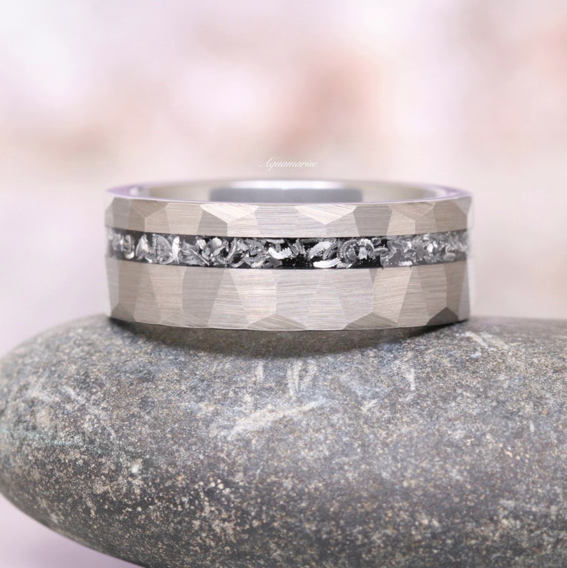 Pu&ograve; includere: Un anello in metallo color argento con una superficie esterna sfaccettata e martellata e un intarsio orizzontale centrale riempito di frammenti scuri, testurizzati e dall'aspetto metallico. La parola Aquamarise &egrave; scritta in piccolo corsivo chiaro.