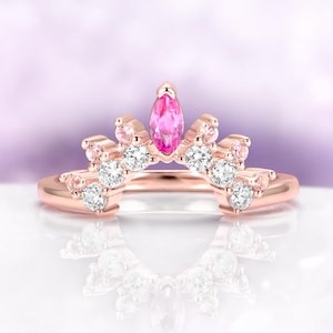 May include: A rose gold ring with a crown design. The ring features a marquise-cut pink gemstone at the center, surrounded by smaller white and pink gemstones. The ring's band curves to create the crown shape.
