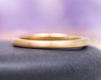 May include: A close-up shot of a gold wedding band with a brushed finish. The ring is set against a blurred purple and white background. The ring is resting on a black stone.