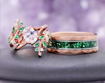 May include: A rose gold engagement ring featuring a central oval clear stone surrounded by leaf-shaped accents with small green and clear stones, paired with a rose gold band featuring a textured surface and a central inlay of crushed green stone.