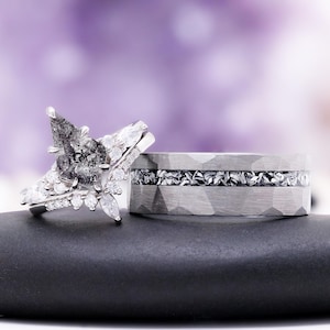 Pu&ograve; includere: Un set di anelli composto da un anello di fidanzamento con diamante a goccia sale e pepe e piccole pietre bianche, abbinato a una fascia larga argentata martellata con un inserto centrale di frammenti metallici scuri e testurizzati.
