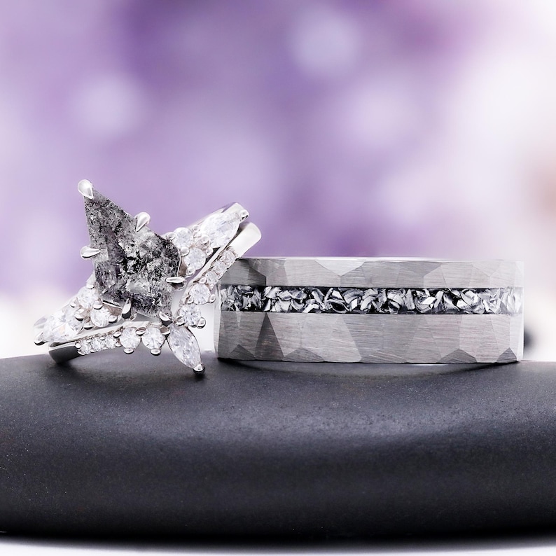 Pu&ograve; includere: Un set di anelli composto da un anello di fidanzamento con diamante a goccia sale e pepe e piccole pietre bianche, abbinato a una fascia larga argentata martellata con un inserto centrale di frammenti metallici scuri e testurizzati.