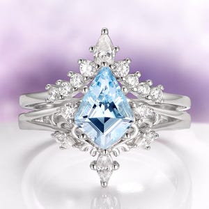 May include: A silver engagement ring set with a large, light blue, kite-shaped gemstone, surrounded by smaller clear stones. The ring has a delicate, ornate design with a matching band. The setting is on a white surface with a purple background.