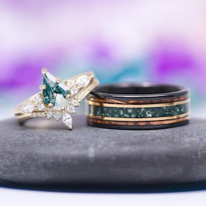 May include: A pair of wedding bands. The engagement ring features a pear-shaped green moss agate gemstone surrounded by smaller diamonds set in a gold band. The wedding band is made of black metal with green and brown inlay.