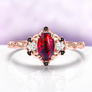 May include: Rose gold ring featuring an oval, fiery red opal center stone. The ring is accented with small white and black gemstones, set in an ornate, filigree design. The band has a delicate, textured pattern.