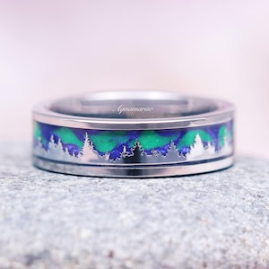 May include: A silver ring with a blue and green inlay and a silver silhouette of a forest. The ring is on a gray rock.