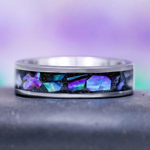 May include: A silver ring with a band inlaid with iridescent abalone shell fragments in shades of blue, purple, and green. The ring has a polished, metallic finish and a dark background for the abalone. The ring is a piece of jewelry.