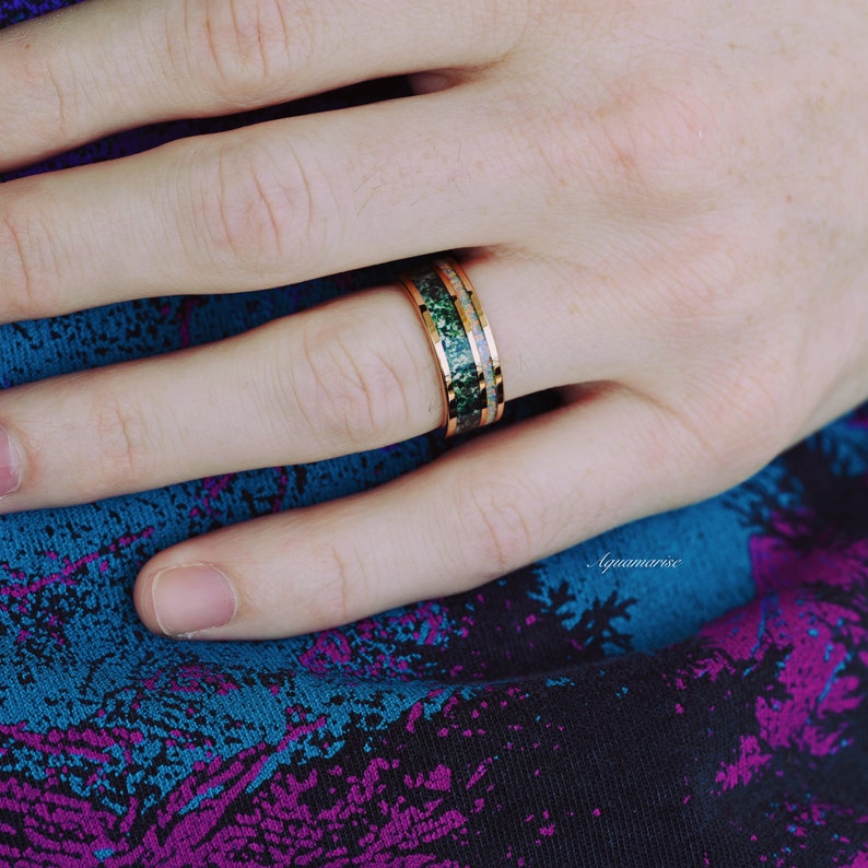 May include: A metallic ring featuring a wide band with a polished rose gold finish. The centre of the band contains a textured inlay of dark green crushed stone flanked by two thin strips of iridescent white opal material on either side.