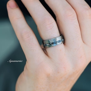 Pu&ograve; includere: Un anello in metallo grigio scuro con una superficie martellata e sfaccettata, caratterizzato da un canale orizzontale centrale intarsiato con piccole pietre blu scintillanti. La parola Aquamarise &egrave; scritta in bianco sul lato sinistro.