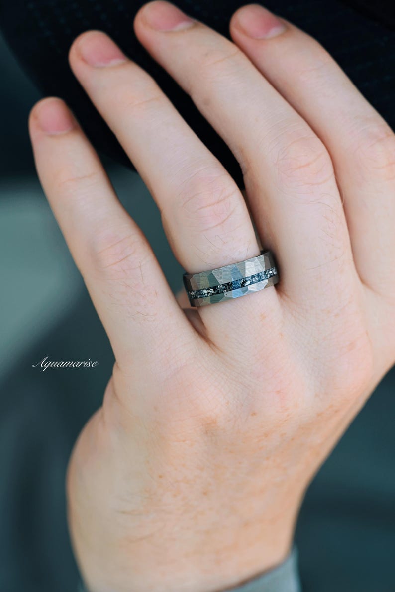 Pu&ograve; includere: Un anello in metallo grigio scuro con una superficie martellata e sfaccettata, caratterizzato da un canale orizzontale centrale intarsiato con piccole pietre blu scintillanti. La parola Aquamarise &egrave; scritta in bianco sul lato sinistro.