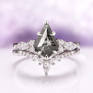 Pu&ograve; includere: Un anello in metallo argentato con una pietra centrale a forma di aquilone, di tipo sale e pepe, fissata da sei griffe. La fascia &egrave; decorata con piccole pietre chiare, rotonde e a taglio marquise, disposte a chevron attorno alla pietra.