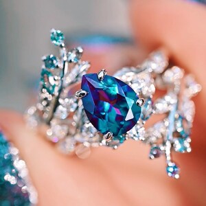 Op de afbeelding: Een zilveren ring met een peervormige blauwe edelsteen omringd door kleinere heldere edelstenen. De ring heeft een delicaat, bladerig ontwerp.