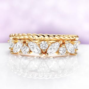 May include: Two gold rings. The top ring has a twisted rope design. The bottom ring is adorned with marquise-cut and round-cut clear gemstones. The rings are set against a soft purple background.