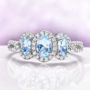May include: A silver ring featuring three oval, light blue gemstones, each surrounded by a halo of small, clear stones. The ring's band has a twisted design with additional small stones.