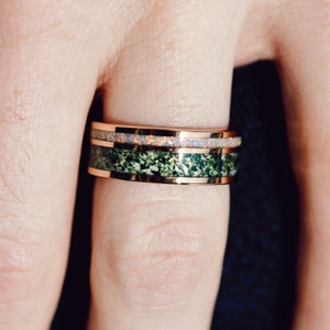 May include: A rose gold band ring featuring two distinct horizontal inlays. The top inlay is a thin strip of iridescent white opal, while the wider bottom inlay consists of a textured, dark green moss-like mineral material.