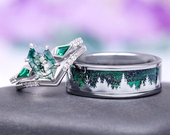 May include: A silver wedding ring set featuring a unique design. The engagement ring has a large, green gemstone and smaller green accent stones. The wedding band has a forest scene with green trees and a starry night design.