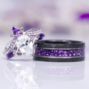 May include: A pair of wedding bands with a purple theme. The engagement ring features a pear-shaped diamond surrounded by a floral design with purple gemstones. The wedding band is black with a band of purple gemstones.