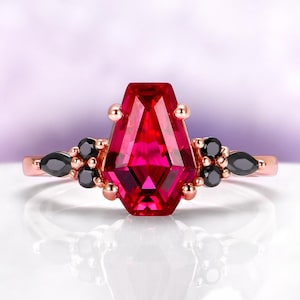 May include: Rose gold ring featuring a large, faceted, red gemstone in a unique hexagonal shape. The ring is accented with small black gemstones and gold spheres on the band, creating a striking contrast. The ring is set against a soft, blurred background.