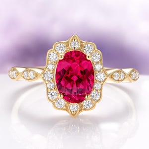 May include: A gold ring featuring a large, oval-cut, ruby-red gemstone surrounded by a halo of small, round, white diamonds. The ring's band has a delicate, ornate design with additional small diamonds.