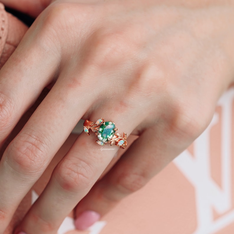 May include: A rose gold ring featuring a central oval-cut green moss agate stone flanked by small marquise-cut opals. The band is designed with a detailed leaf and vine motif that extends along the sides, encircling the finger with metal foliage.
