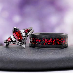 May include: A set of two black metal rings. One ring features a central kite-shaped red gemstone flanked by smaller red stones and clear round accent stones. The second ring is a wide band with a hammered texture and a central inlay of red fragments.