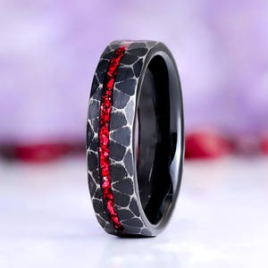 May include: A black ring featuring a hammered texture with a central inlay of crushed red material. The band has a smooth, polished interior and a dark, matte exterior finish with a distinct pattern of dark gray and black geometric shapes.