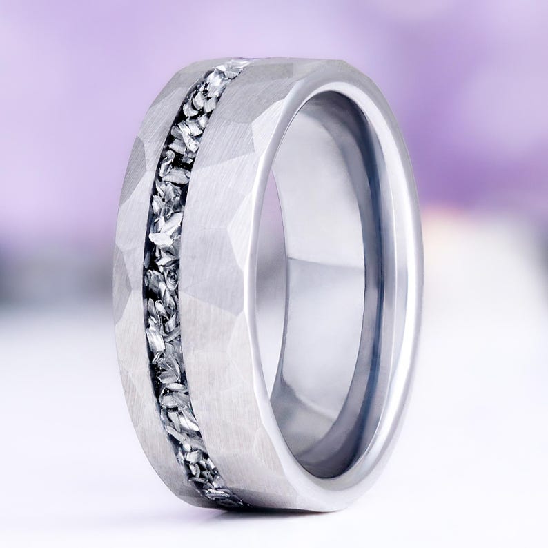 Pu&ograve; includere: Un anello color argento caratterizzato da una superficie esterna martellata e testurizzata con un inserto centrale composto da piccoli frammenti metallici frastagliati. La fascia ha un profilo largo con finitura interna lucida e aspetto robusto.