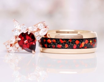 May include: A rose gold engagement ring featuring a pear-cut deep red garnet stone surrounded by marquise and round white stones, paired with a rose gold band inlaid with a black glittery material and scattered orange-red stone fragments.