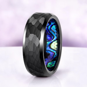 May include: A black ring with a hammered texture on the outside and a vibrant, iridescent abalone shell inlay. The ring's interior displays shades of blue, green, and purple. The ring is a bold piece of jewelry.