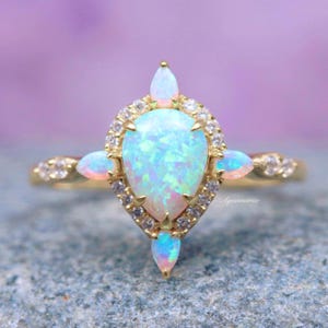 May include: A pear-shaped opal ring with a halo of smaller stones. The ring is set in gold and has a delicate design. The opal is a beautiful iridescent blue and green.