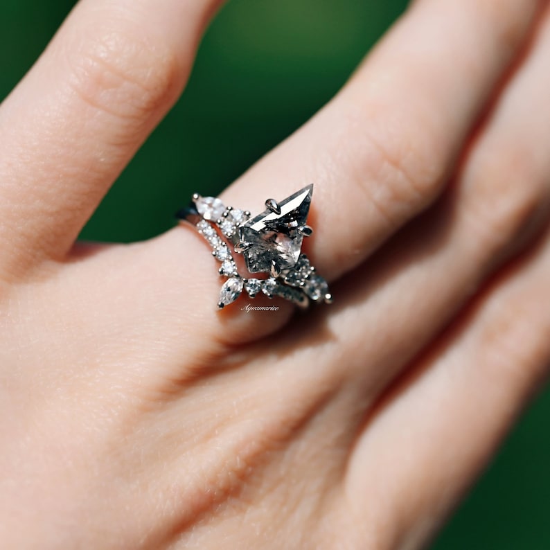 Pu&ograve; includere: Un set di anelli color argento con una pietra centrale di diamante sale e pepe a forma di aquilone con incastonatura a griffe. La fascia &egrave; decorata con piccole pietre chiare a taglio marquise e rotondo disposte a chevron sovrapposto.