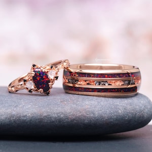May include: Rose gold wedding rings set on a gray stone. The engagement ring features a dark opal with black and white accents. The matching band has a dark red and black inlay with copper-colored flecks.